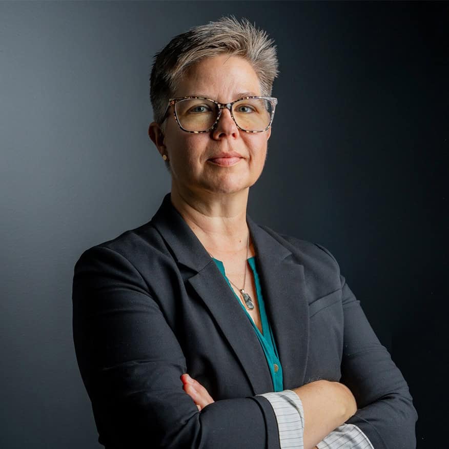 A person with short hair and glasses stands confidently with arms crossed, wearing a dark blazer and a teal shirt. The background is a plain, dark gray wall.
