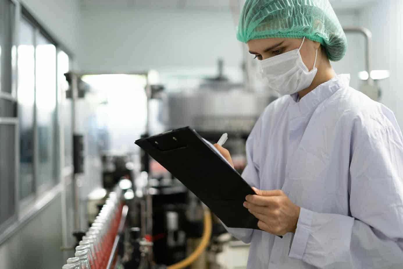 A person in protective clothing, a hair net, and a face mask writes on a clipboard while inspecting bottles on a production line in a laboratory or factory setting.
