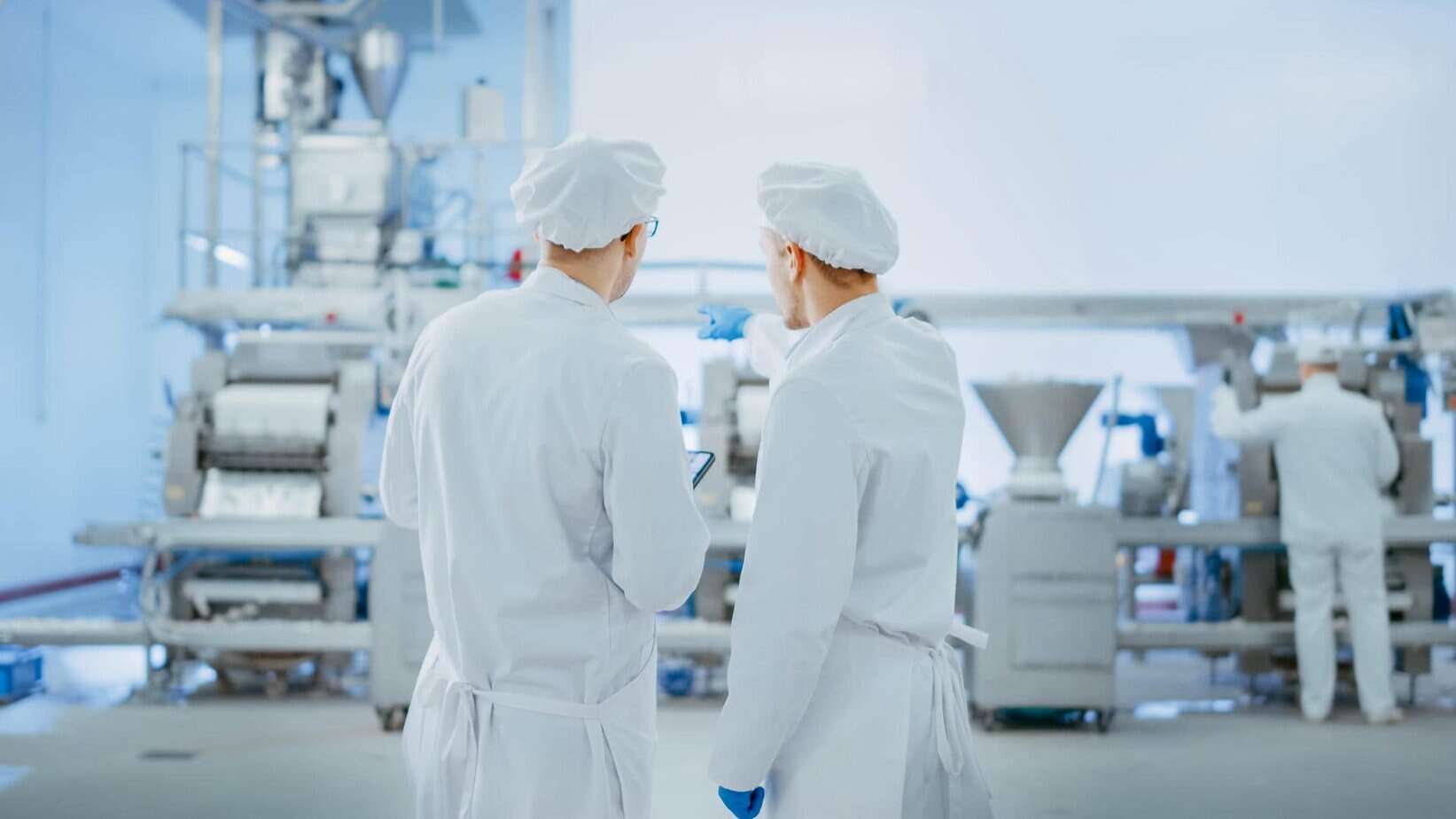 Two people in white lab coats, hairnets, and gloves stand in a bright, clean food processing facility, facing industrial machinery, while a third person works in the background.