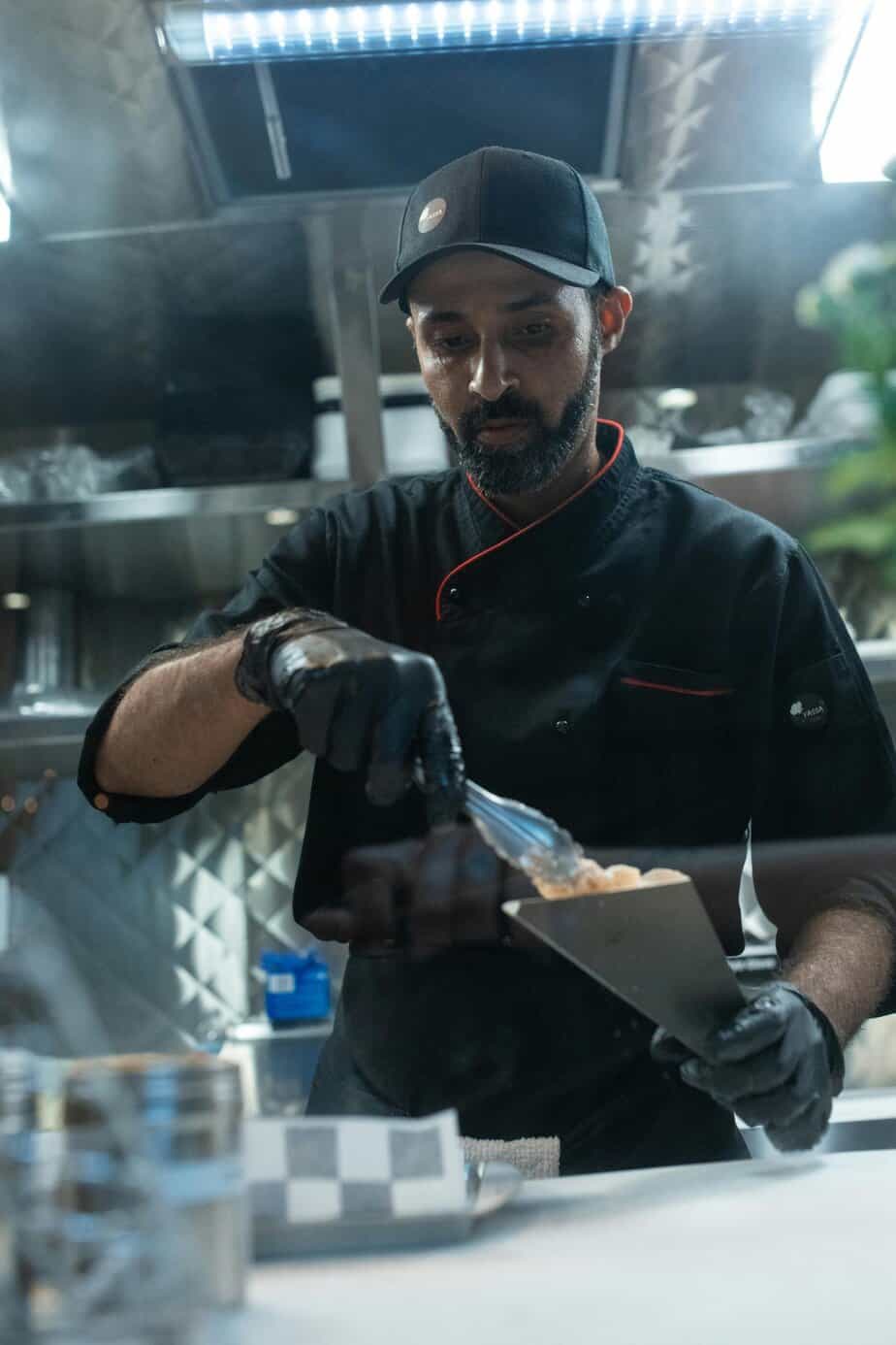 A chef wearing a black uniform and cap is plating food with tongs in a professional kitchen, focused on his task.