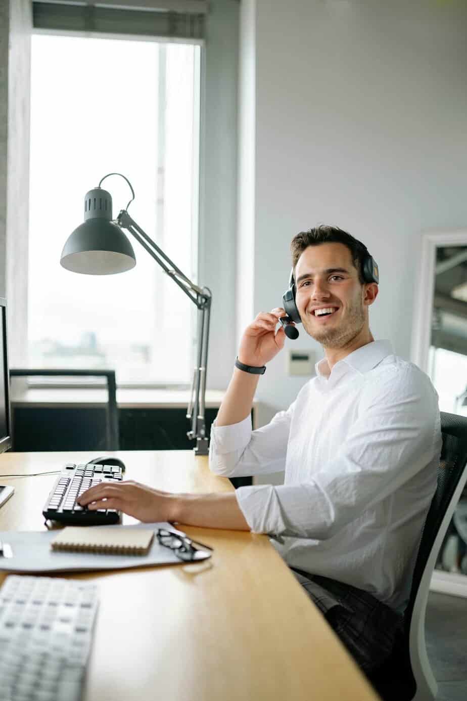 A smiling man wearing a white shirt and headset sits at a desk, using a computer keyboard and mouse. He is looking up, possibly engaged in a phone call or video meeting in a modern office setting.