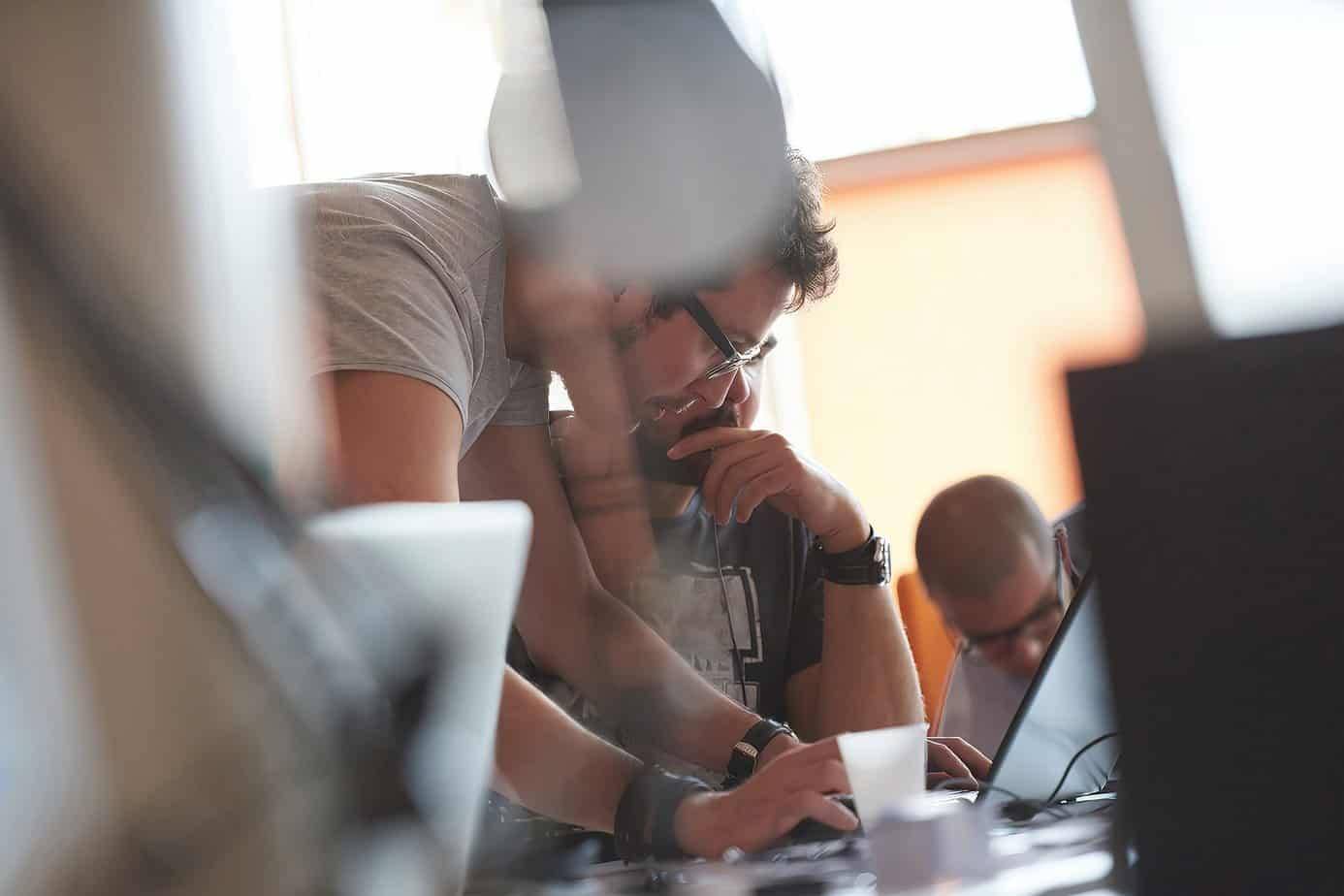 Two people closely collaborate while looking at a laptop screen, with one person pointing at the screen and the other resting his chin on his hand. Another person is blurred in the background.